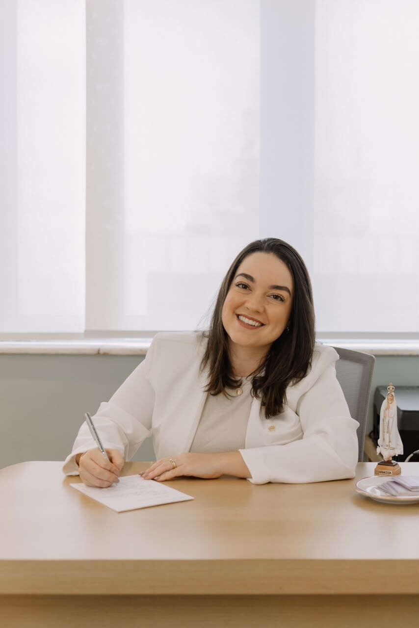 Dra. Thaís Elmôr Semprine escrevendo à mesa do consultório, de blazer branco